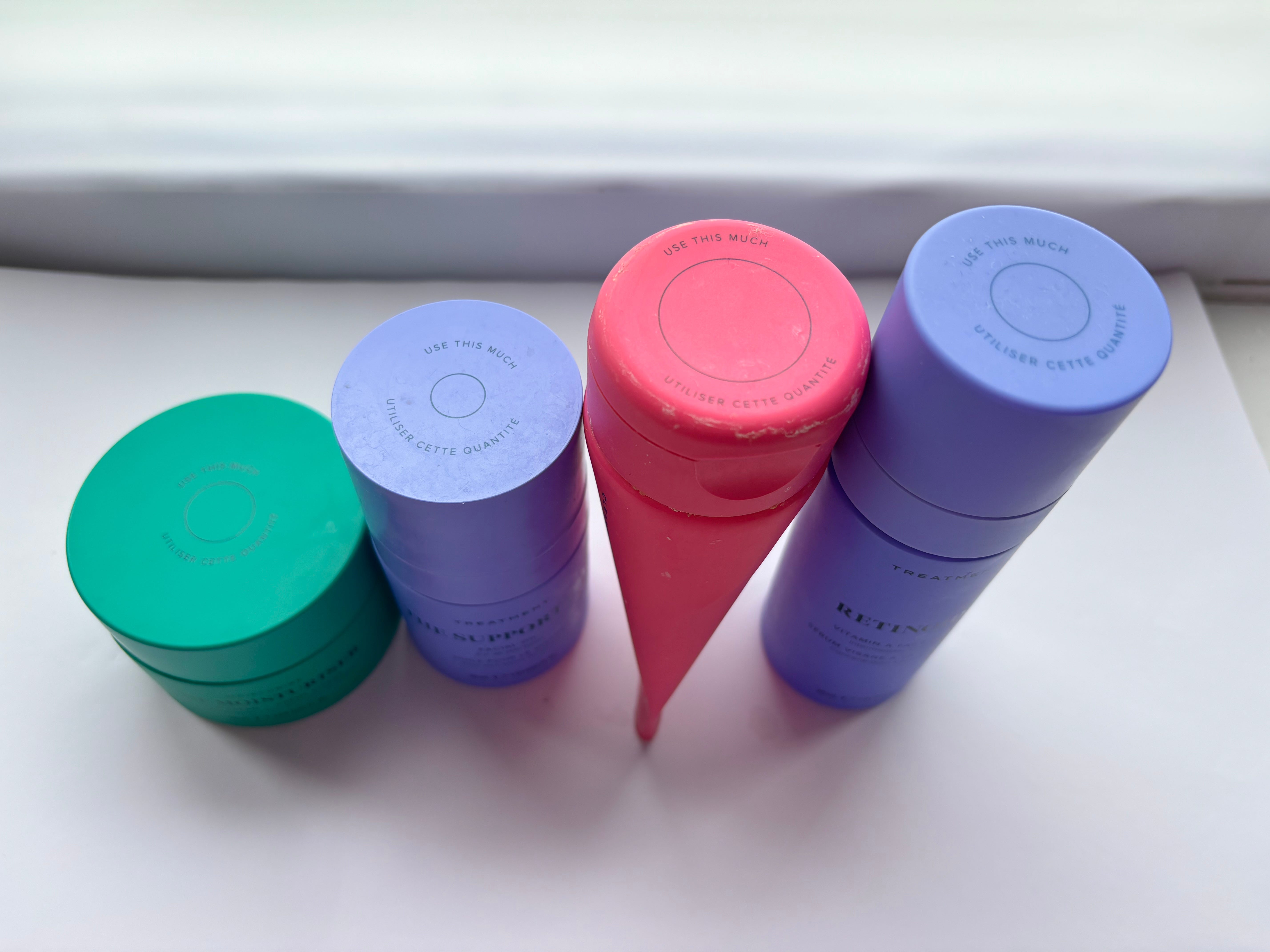 Three Skin Rocks products lined up with the caps of the products showing. From left to right, there is a green jar of face cream, then a purple bottle of skin oil, then a hot pink tube of face wash and finally a bottle of retinol. Each product has a circle on the lid that says "use this much" to represent how much of the product you should use on your face.