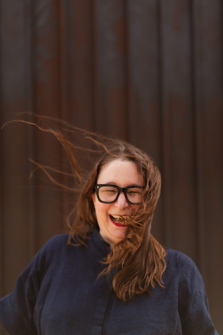 A white woman is laughing as her coppery hair is whipped up by the wind she is wearing large glasses and a navy blue blouse